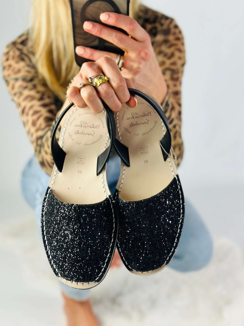 A person holds up a pair of black glitter  flats with black leather straps and beige jute soles. The person is wearing a leopard print top and light blue jeans, 