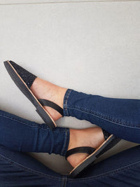 A top-down view of a person wearing black pointed-toe flat sandals with black ankle straps and glittery black toes, paired with blue jeans