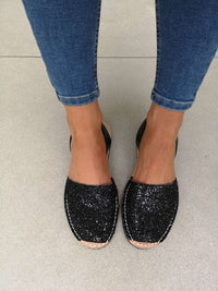 Black glitter slip-on espadrille-style flats worn with blue skinny jeans, shown from a top-down view on a tiled floor.
