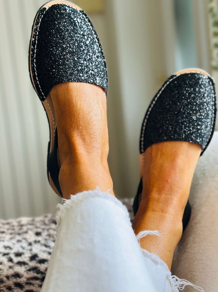 A close-up lifestyle shot of a person wearing black glitter slip-on avarca flats with white contrast stitching, paired with white frayed-hem jeans.