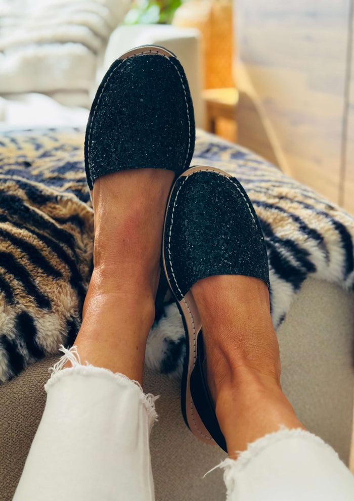 A person wearing black glitter slip-on avarcas with white frayed-hem jeans, sitting on a couch with a leopard-print throw blanket.