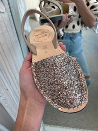 A person holds a glittery sandal covered in rose gold glitter in front of a mirror.