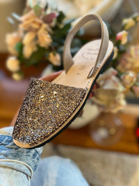 A person holds a glittery flat sandal with a beige slingback strap, shown against a soft-focus background of flowers and a wooden surface.
