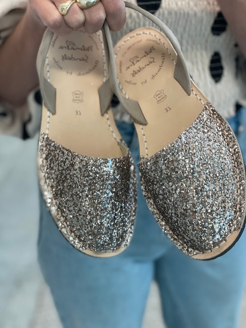 A person holds up a pair of women's flat sandals covered in glitter, with beige slingback straps. The brand name 'Palmaira Sandals' is visible on the insole.
