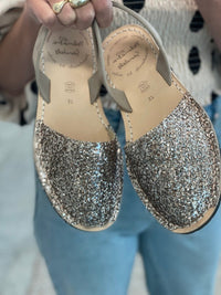 A person holds up a pair of women's flat sandals covered in glitter, with beige slingback straps. The brand name 'Palmaira Sandals' is visible on the insole.
