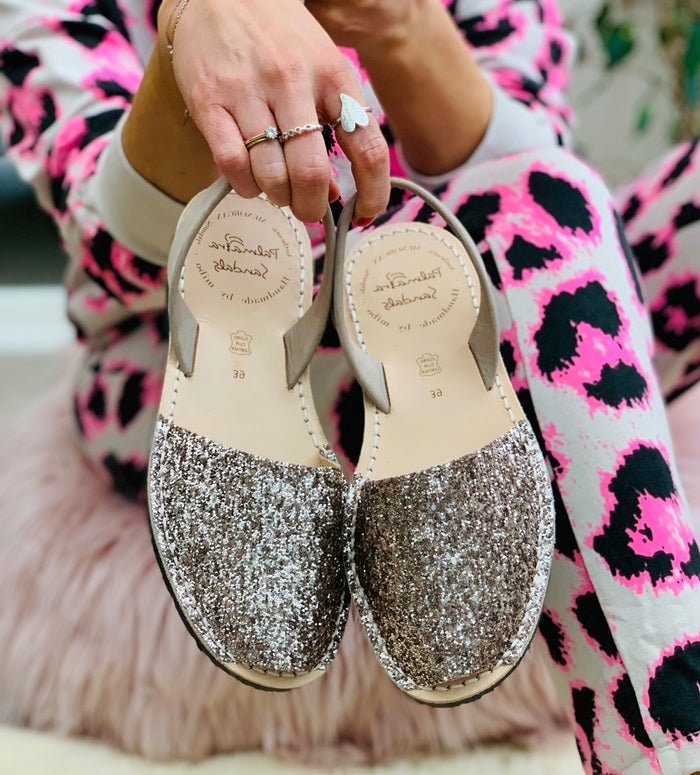 A person holds a pair of women's flat sandals with glittery silver and champagne beige uppers and thin beige straps. 