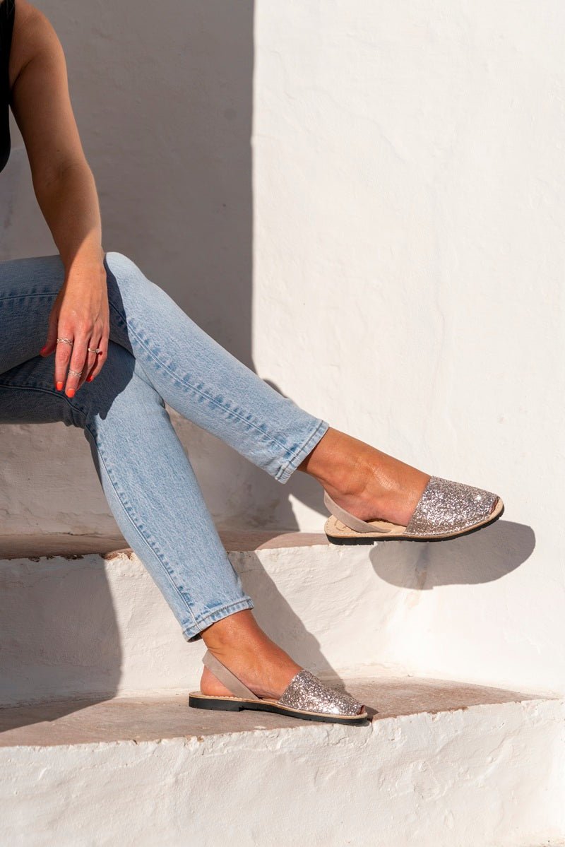 A person seated on white outdoor steps wearing light blue jeans and glitter slingback sandals with black soles, against a white stucco wall.