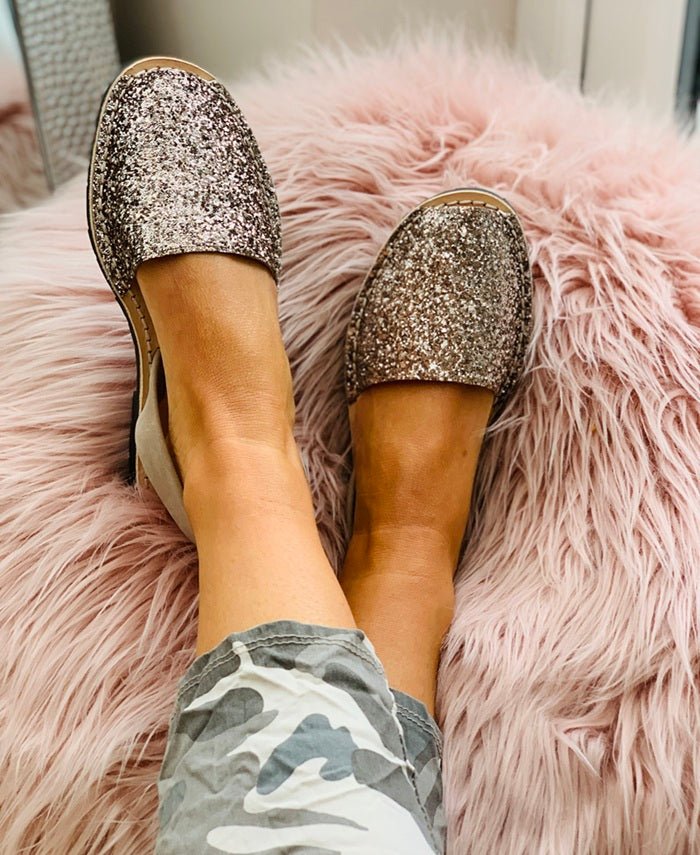 A person wearing glittery slip-on sandals shoes with a metallic rose-gold finish, sitting on a fluffy pink faux-fur rug.