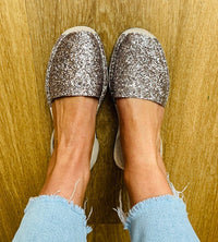 A pair of women's slip-on avarcas covered in sparkling glitter, shown on a person's feet against a wooden floor background.
