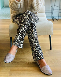 A person seated on a cream upholstered chair wearing black-and-white leopard print pants, and glittery slip-on sandals on a light wood floor.