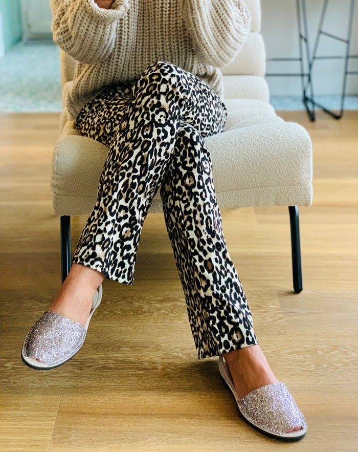 A person seated on a cream upholstered chair wearing black-and-white leopard print pants, and glittery slip-on sandals on a light wood floor.
