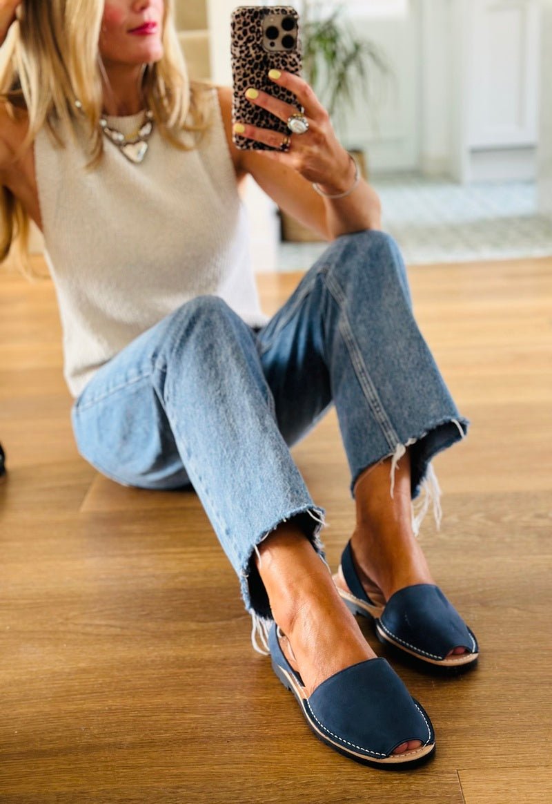 Woman sitting on a wooden floor wearing blue sandals, jeans, and a white top.