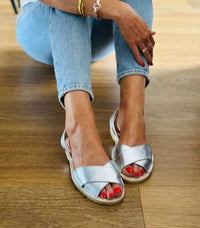 A person wearing metallic silver slide sandals with crisscross straps and a braided jute sole, paired with light blue jeans, sitting on a wooden floor.