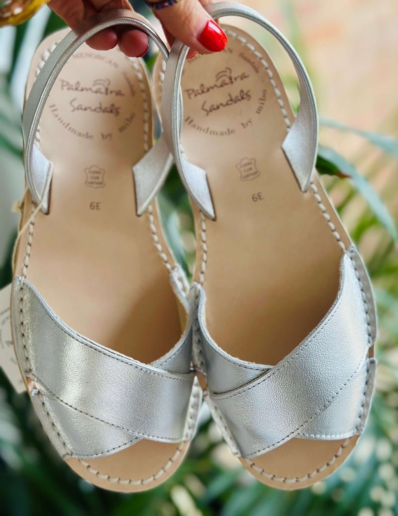 A pair of metallic silver leather slide sandals with crisscross straps and adjustable ankle straps, held by a hand with red nail polish against a blurred background of green foliage.