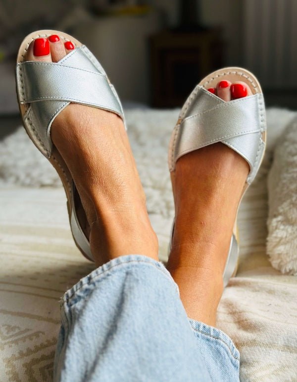 A person wearing metallic silver criss-cross slide sandals with a beige platform sole, shown from the feet up while sitting on a textured cream sofa. The person is wearing light blue jeans and has bright red painted toenails.