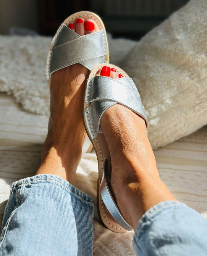 A person wearing metallic silver slide sandals with crisscross straps and a light beige sole, shown in a cozy indoor setting with blue jeans and red toenail polish.