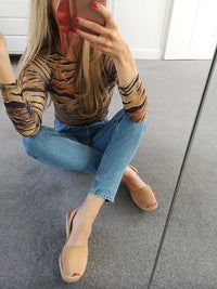 A woman sitting on a grey carpeted floor in front of a mirror, wearing a fitted long-sleeve tiger-print top in brown and black, blue jeans, and beige slingback wedge sandals.