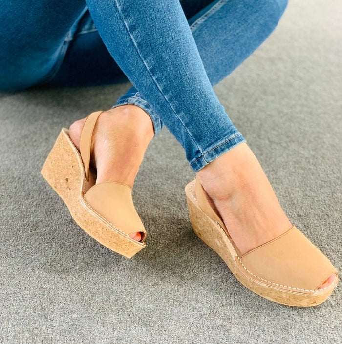 A person wearing tan peep-toe wedge sandals with cork-look soles, paired with blue jeans, sitting on a grey carpeted floor.