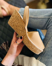 A person holds a tan platform wedge sandal with a cork-textured sole, shown in a close-up lifestyle shot with blue jeans and a hand with black nail polish visible.