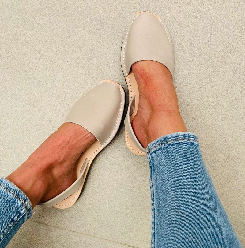 A pair of stone coloured leather slip-on shoes with pointed toes, shown being worn with blue jeans on a neutral concrete floor.