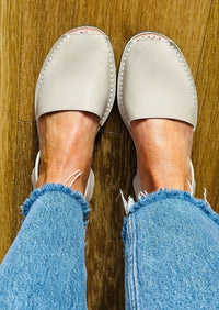A top-down view of a person wearing light stone slip-on sandals with frayed blue jeans, standing on a wooden floor.