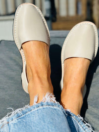A close-up lifestyle shot of a person wearing a pair of light beige slip-on avarcas with frayed blue denim jeans, sitting outdoors.