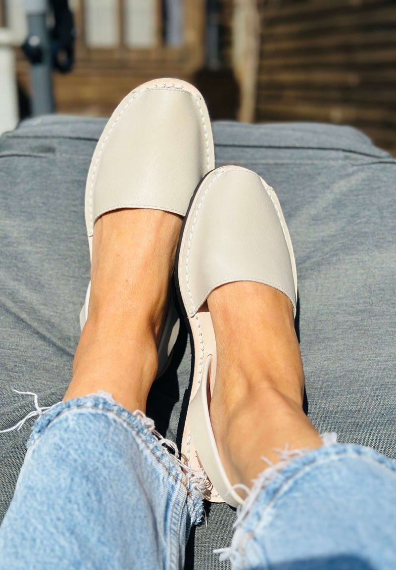 A close-up of a person's feet wearing light beige slip-on avarcas with frayed blue denim jeans, sitting on a couch.
