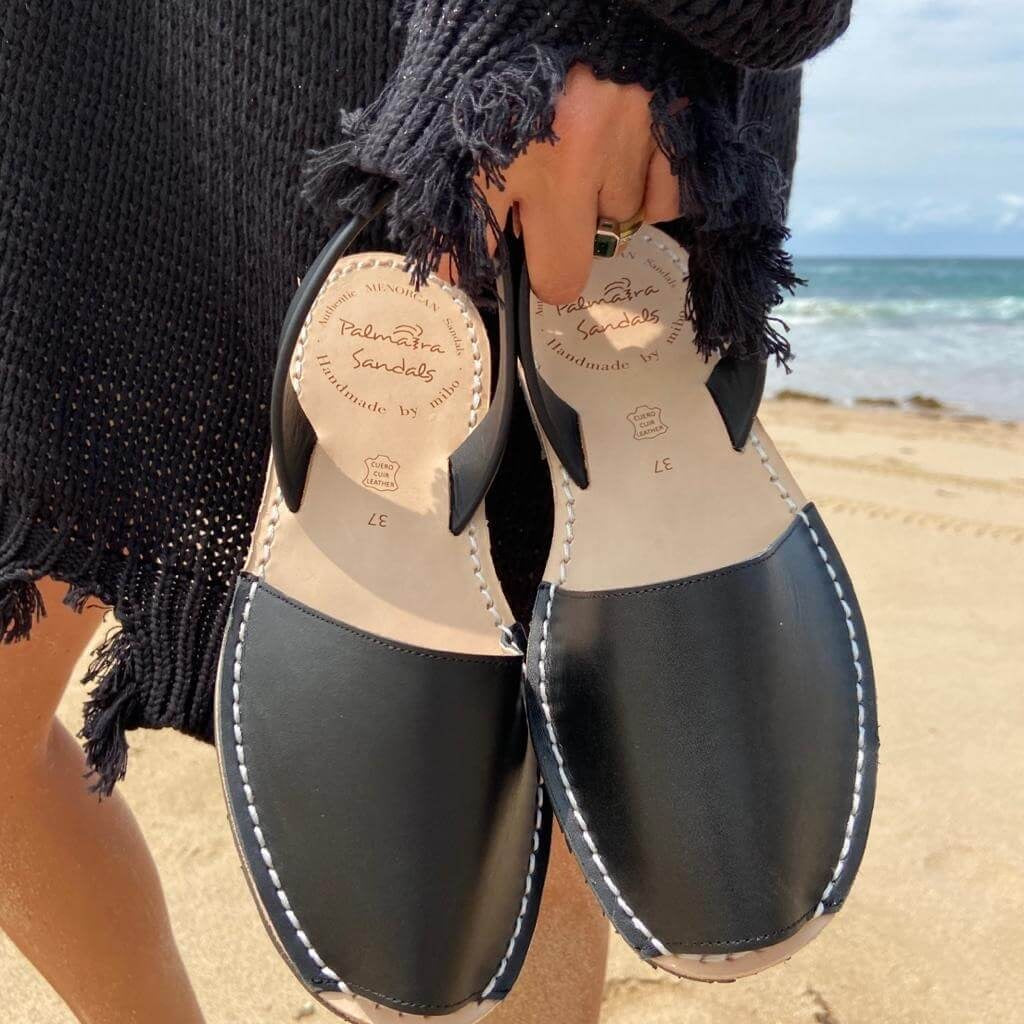 Black sandals with white stitching held by a person on a beach.