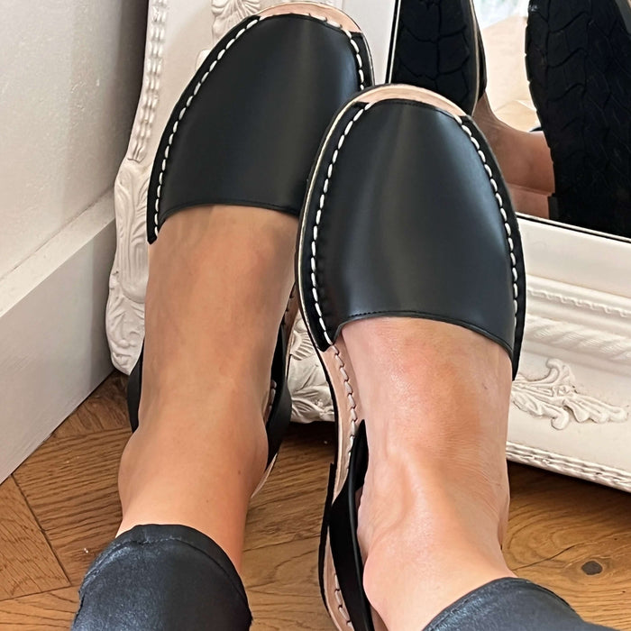 Black leather sandals worn indoors on a wooden floor.