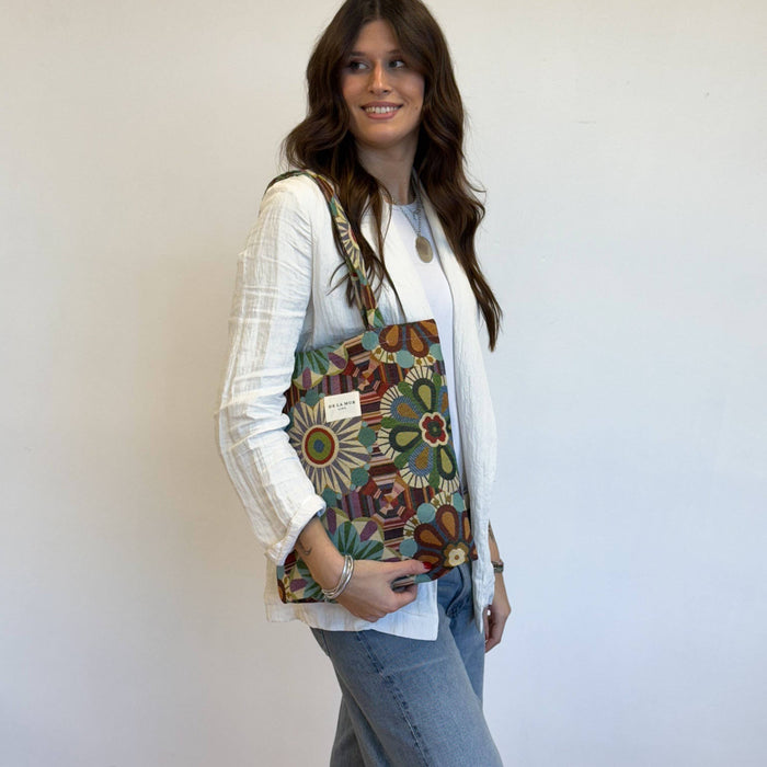 A woman wearing a white cardigan and blue jeans carries a colorful floral tote bag with a patterned strap against a plain white wall.