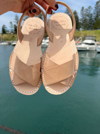 Pair of tan sandals held above a body of water with boats and trees in the background