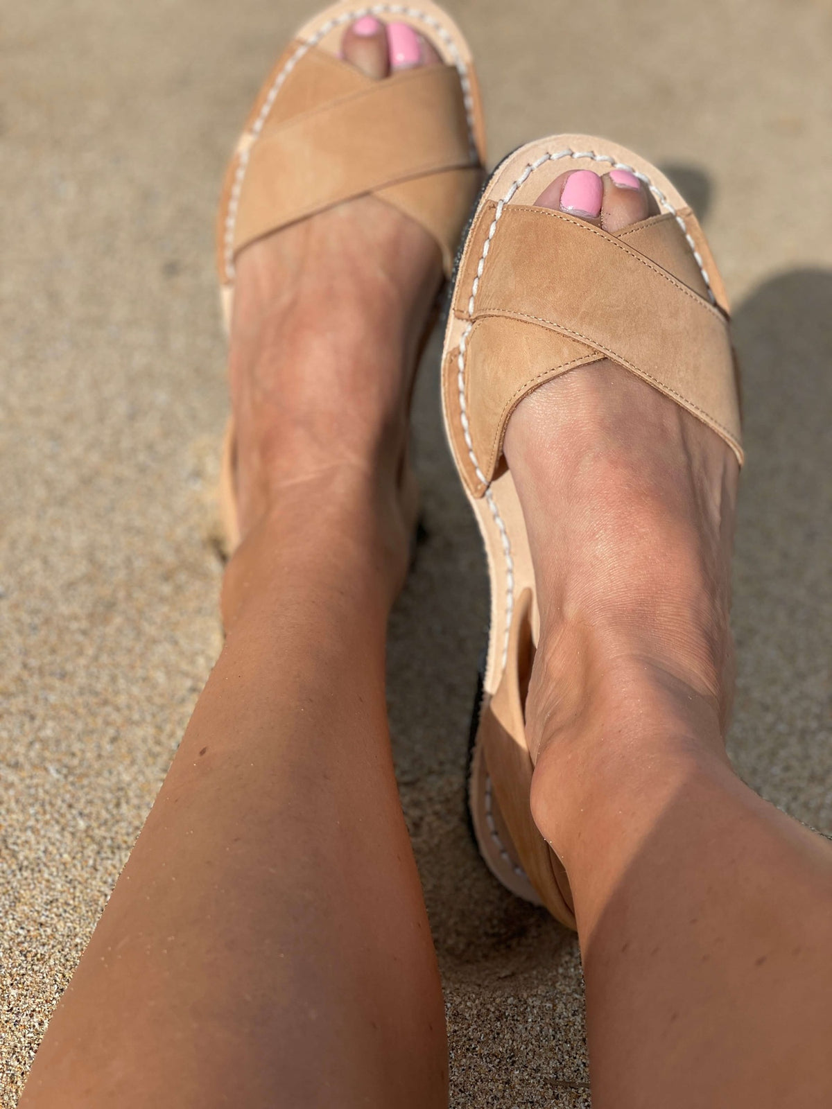 Tan sandals worn on feet with pink nail polish on a sandy surface