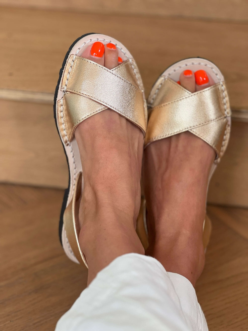 Gold sandals worn on feet with orange nail polish against a wooden floor background