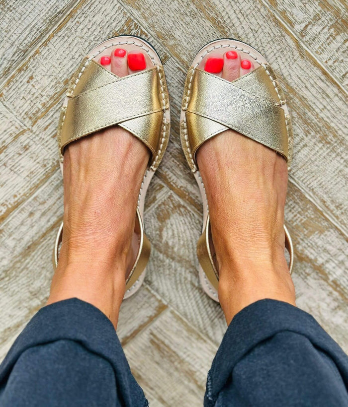 Gold sandals worn on a person's feet with red nail polish, standing on a wooden floor.