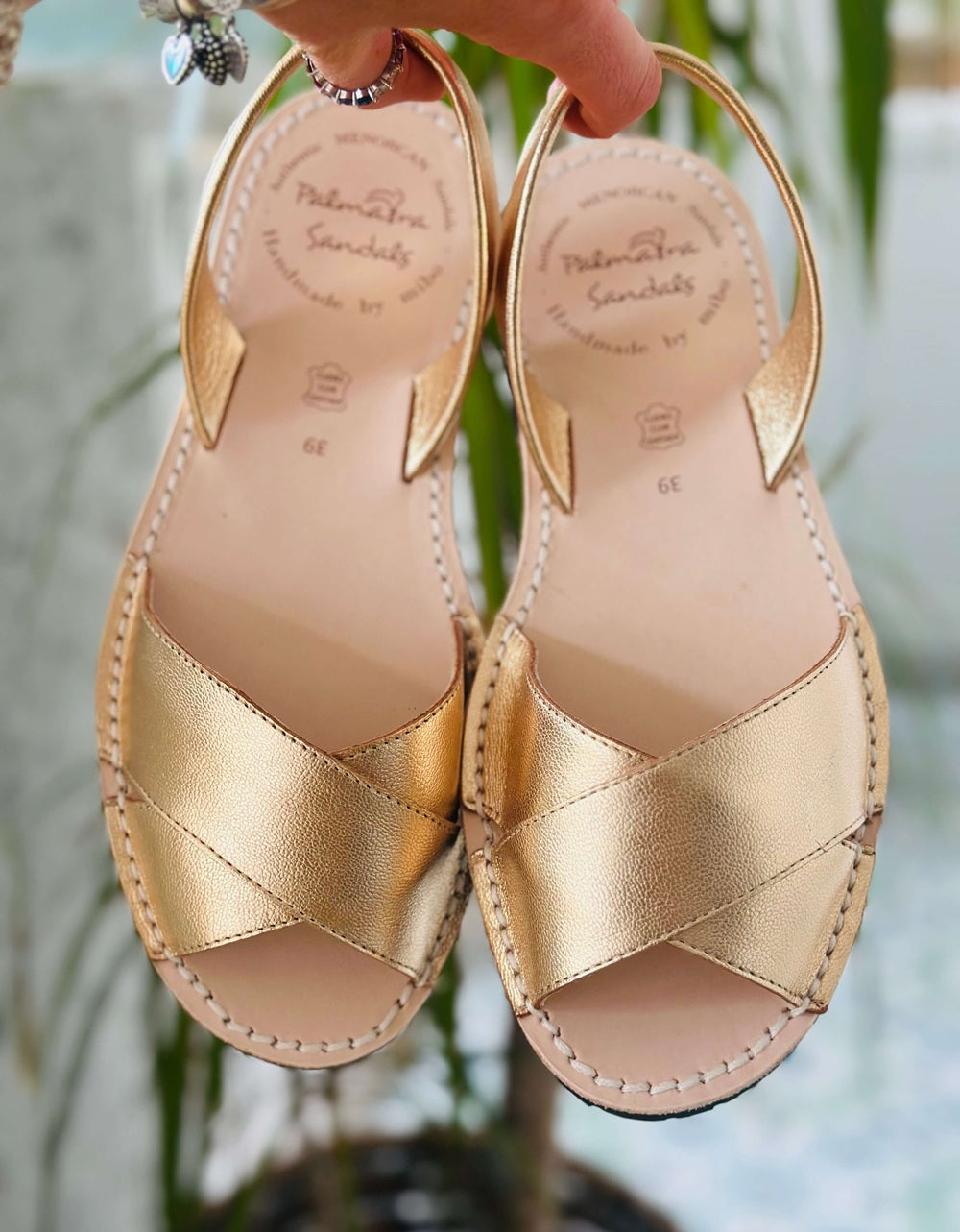 Gold sandals with criss-cross straps held by a hand against a blurred natural background