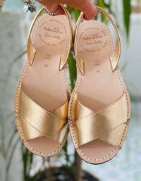Gold sandals with criss-cross straps held by a hand against a blurred natural background