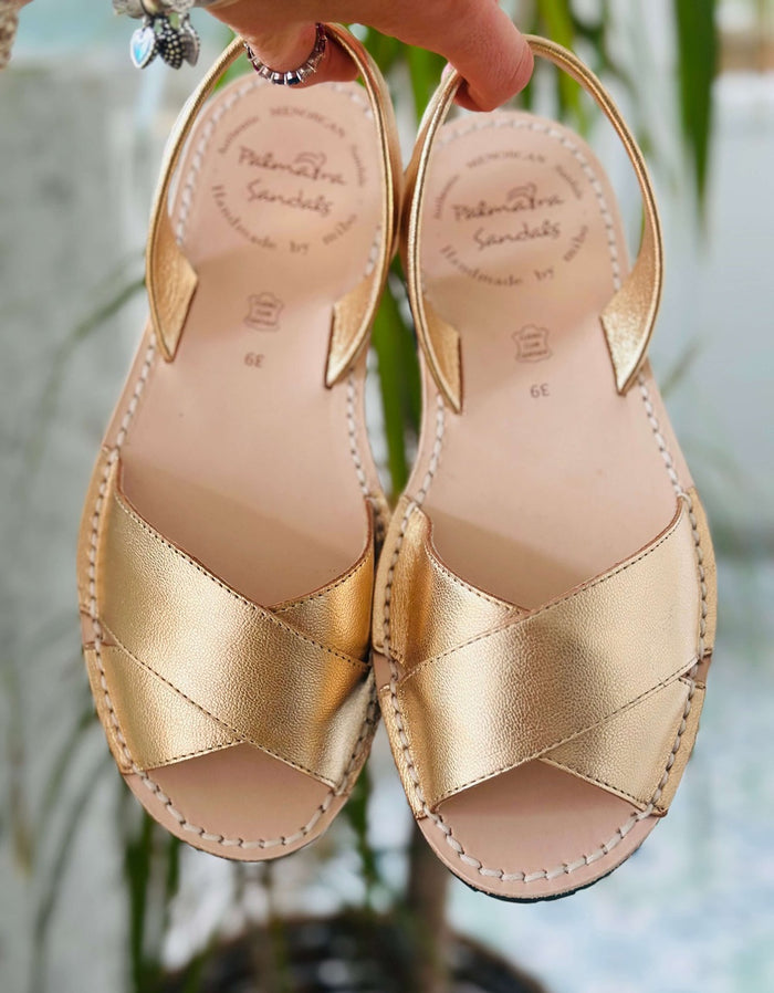 Gold sandals with criss-cross straps held by a hand against a blurred natural background