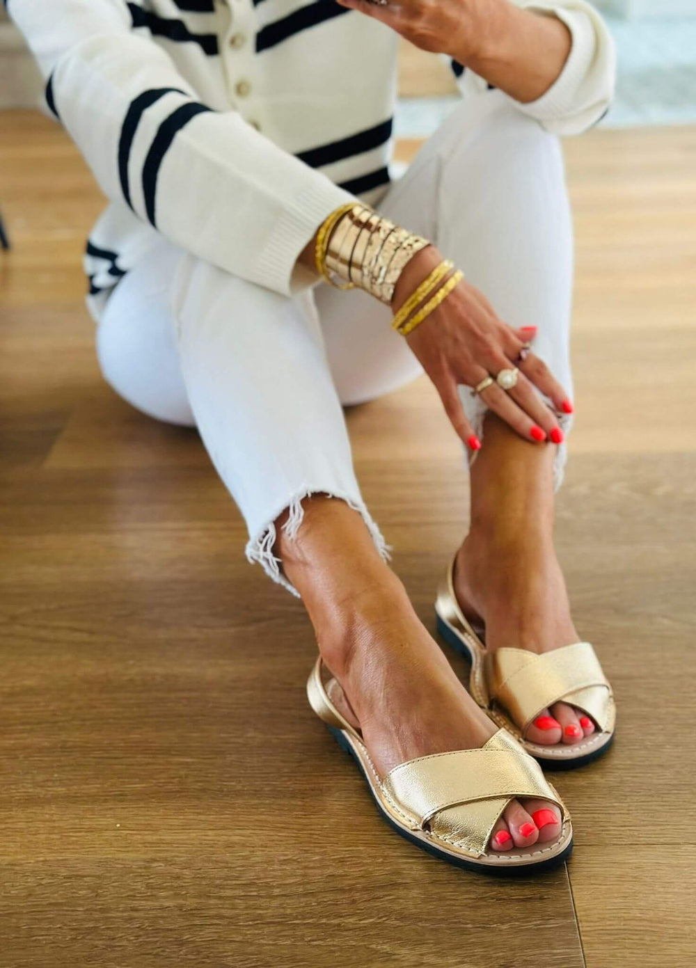 Person wearing gold sandals and white pants sitting on a wooden floor.