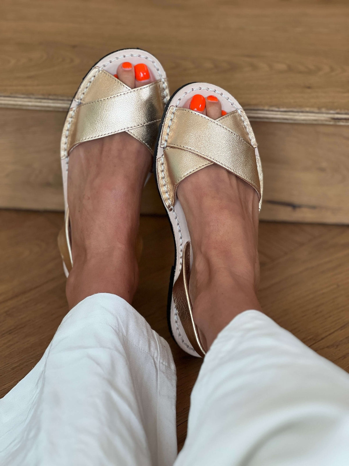 Gold sandals worn with white pants on a wooden floor