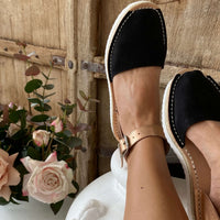 Person wearing black espadrilles with rose gold straps, sitting on a white stool with a wooden background and flowers.