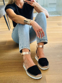 Person wearing black sandals with rose gold straps on a wooden floor