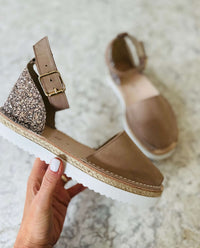 Taupe sandals with glittery backpiece held by a hand on a light background