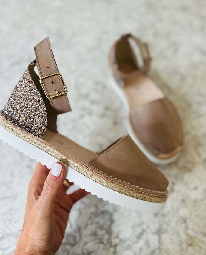 Taupe sandals with glittery backpiece held by a hand on a light background
