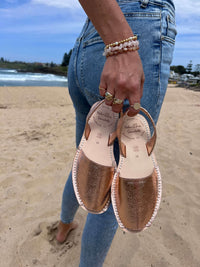 Person holding a pair of sandals on a beach with ocean and sky in the background