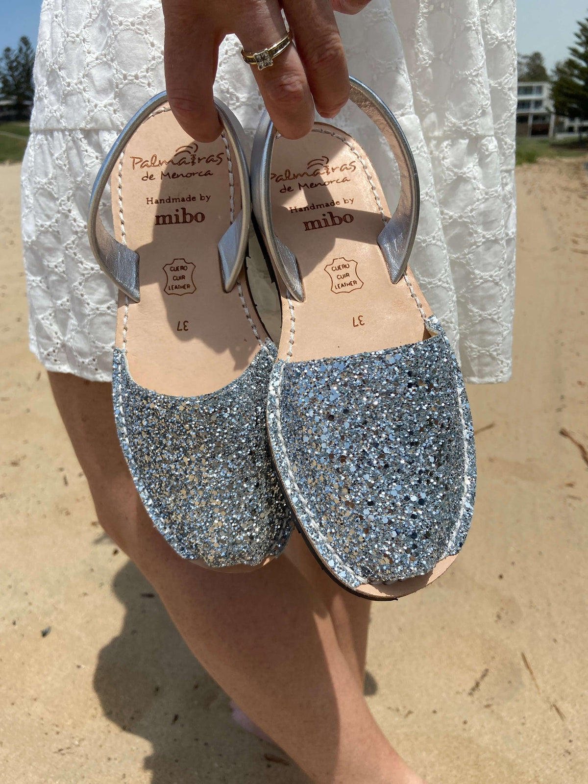 Pair of silver glitter sandals held by a person on a sandy background