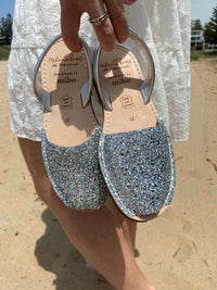 Pair of silver glitter sandals held by a person on a sandy background
