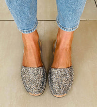 Pair of glittery flats worn with rolled-up jeans on a neutral floor.