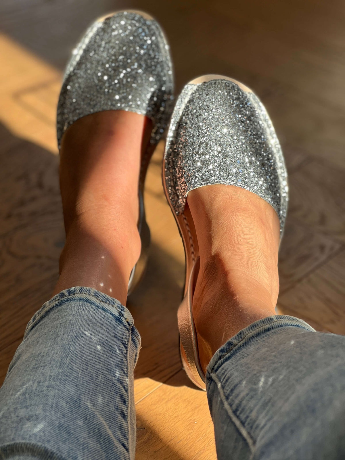 Silver glittery shoes worn with blue jeans on a wooden floor.